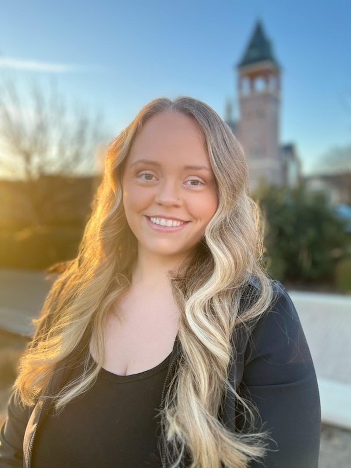 Professional headshot of a smiling woman with long blonde hair, wearing a black blazer, with a blurred clock tower in the background