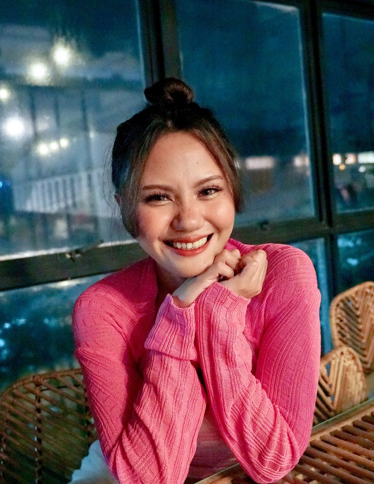 Smiling woman in a pink sweater with her hair in a bun, sitting at a wicker table with warm lighting and a glass window in the background.